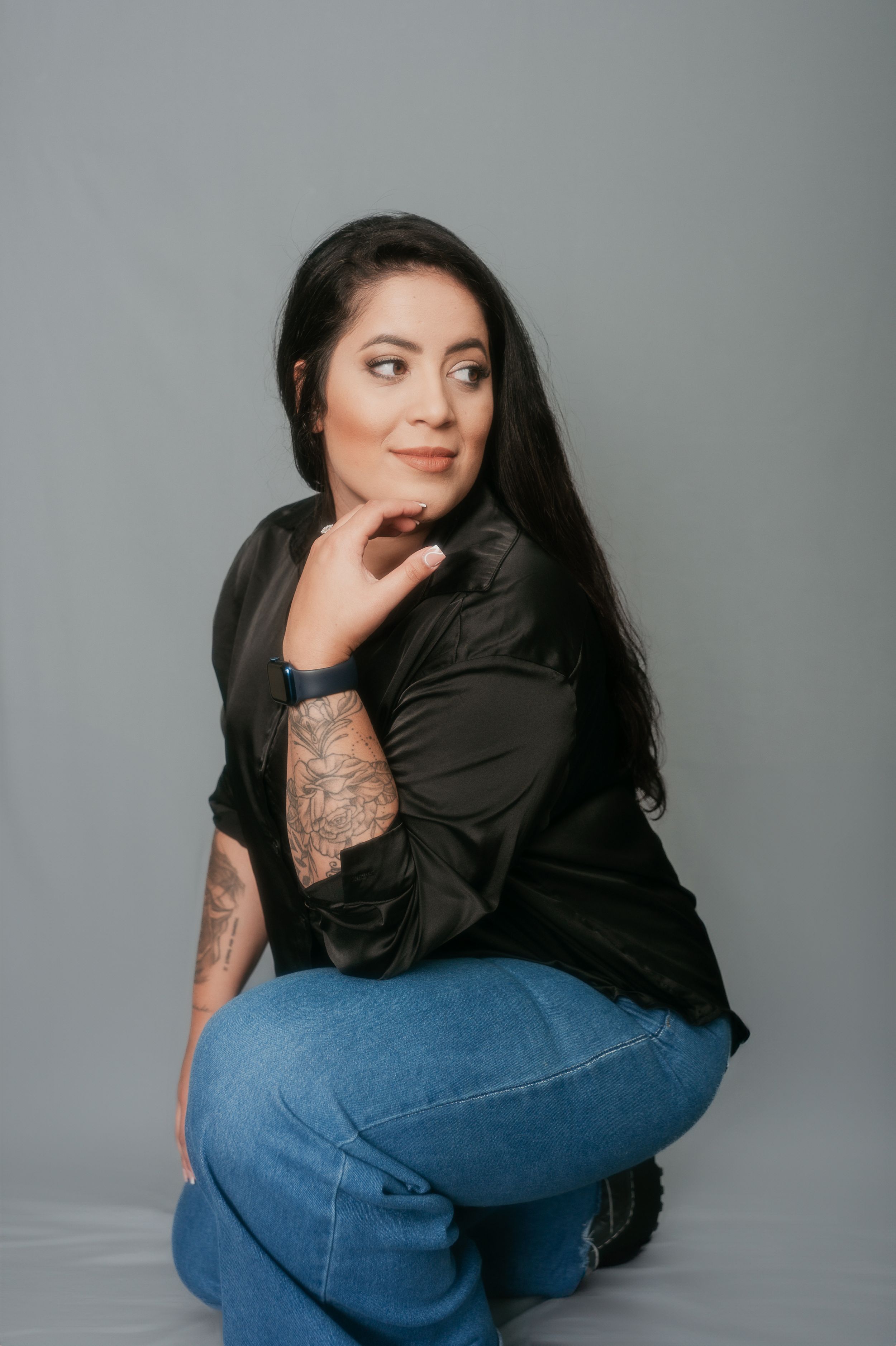 Professional studio portrait of a person in a black button-up shirt and jeans posing against a gray background.