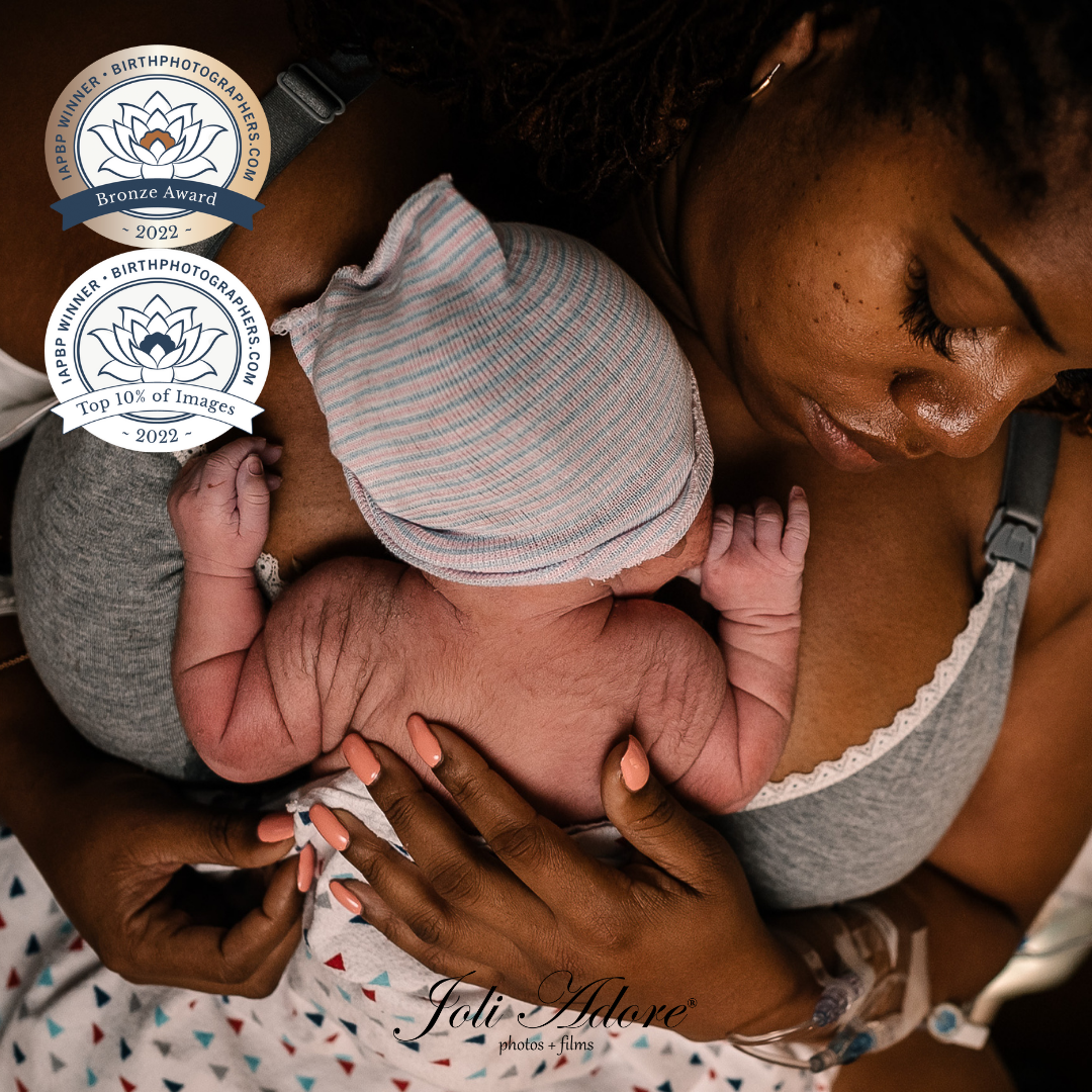 award winning black birth photographer - woman in an gray nursing bra holding a baby wearing a hat