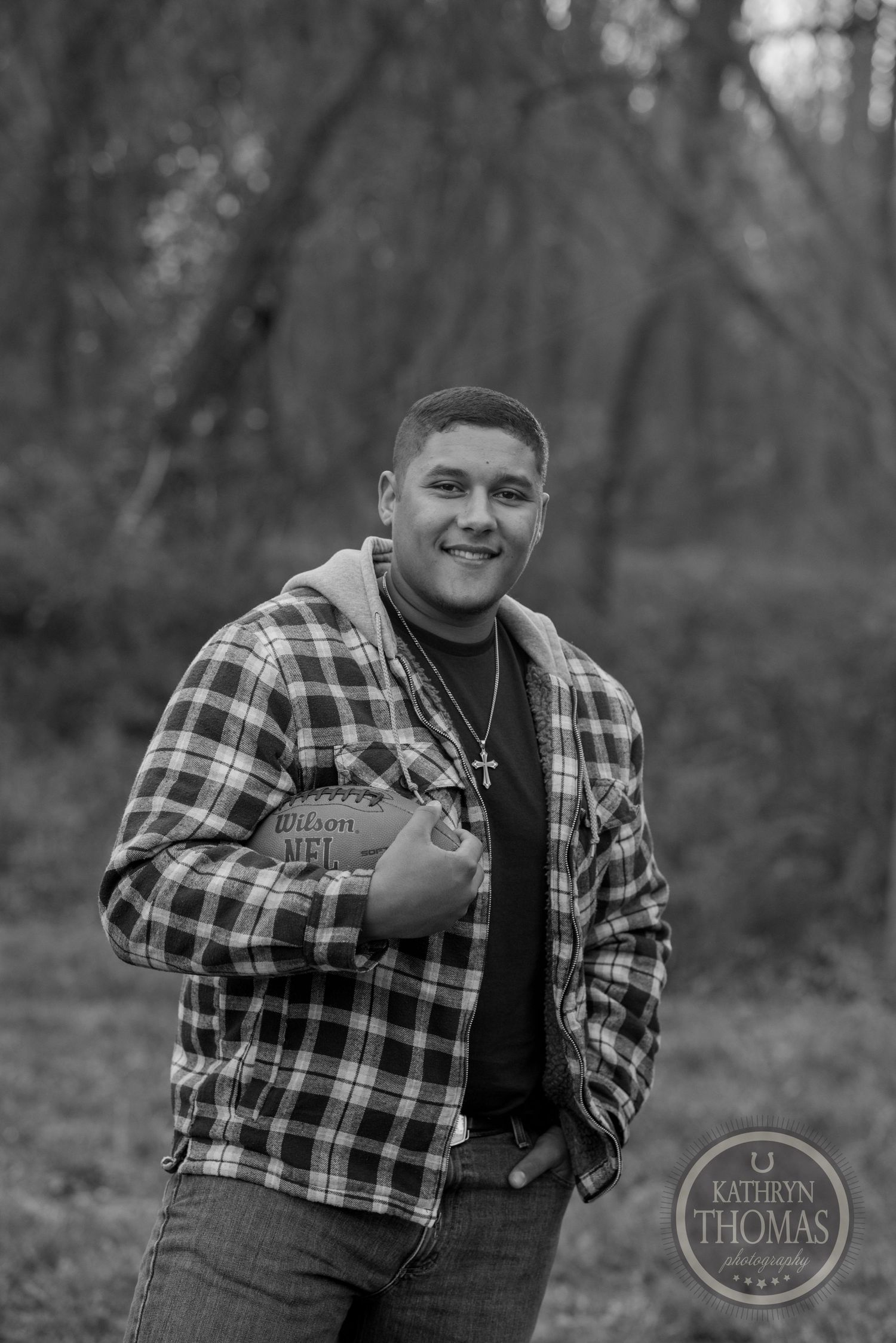 A black and white portrait of someone holding a football while wearing plaid in the woods.
