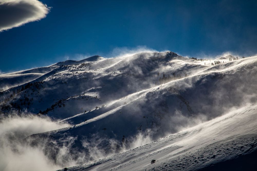 Snow-covered mountain peak with wind-blown snow and clouds creating dramatic atmosphere.