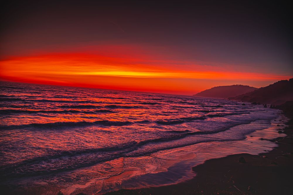 Dramatic red sunset creates vibrant reflections on ocean waves along coastal shore.