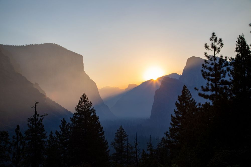 Golden sun peeks through mountain valley with silhouetted pine trees in Yosemite National Park.