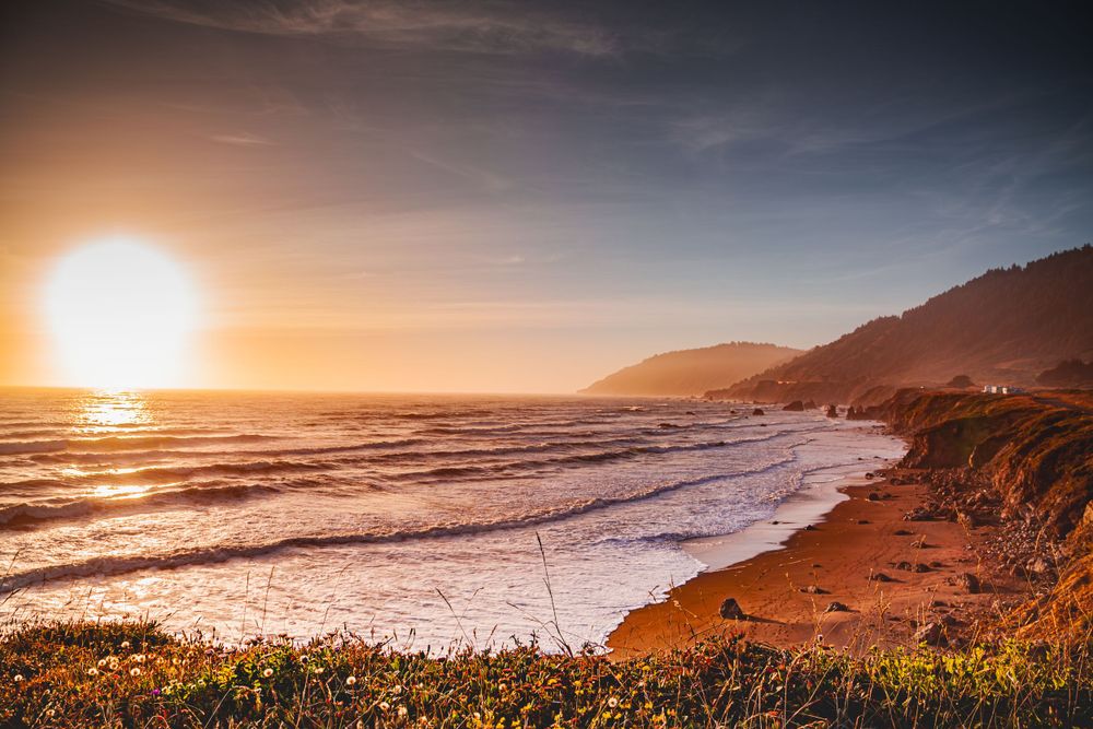 Golden sunset illuminates coastal beach with rolling waves and mountainous backdrop.