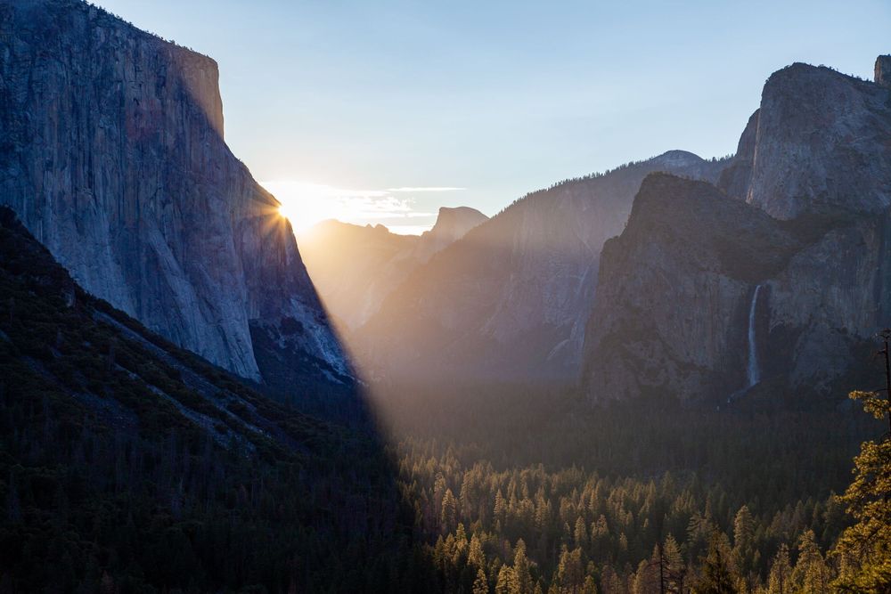 Sunlight beams through Yosemite Valley at sunset, illuminating towering granite cliffs and forest below.