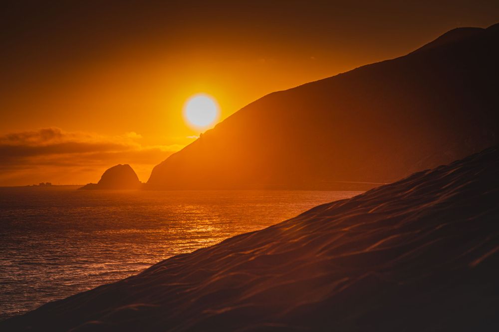 Dramatic sunset silhouetting coastal mountain ridge against orange sky over Pacific Ocean.