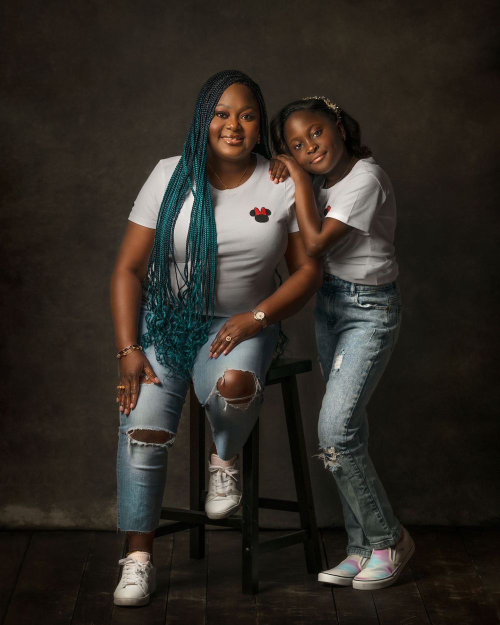 Portrait of mother with beautiful blue braids and her daughter in casual white tops and ripped jeans posing together in portrait studio.
