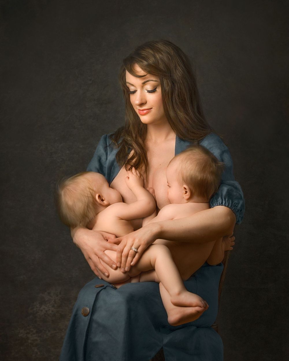 An intimate nurturing moment captured in classic portrait style against a dark background, of a mother breastfeeding her twin babies.
