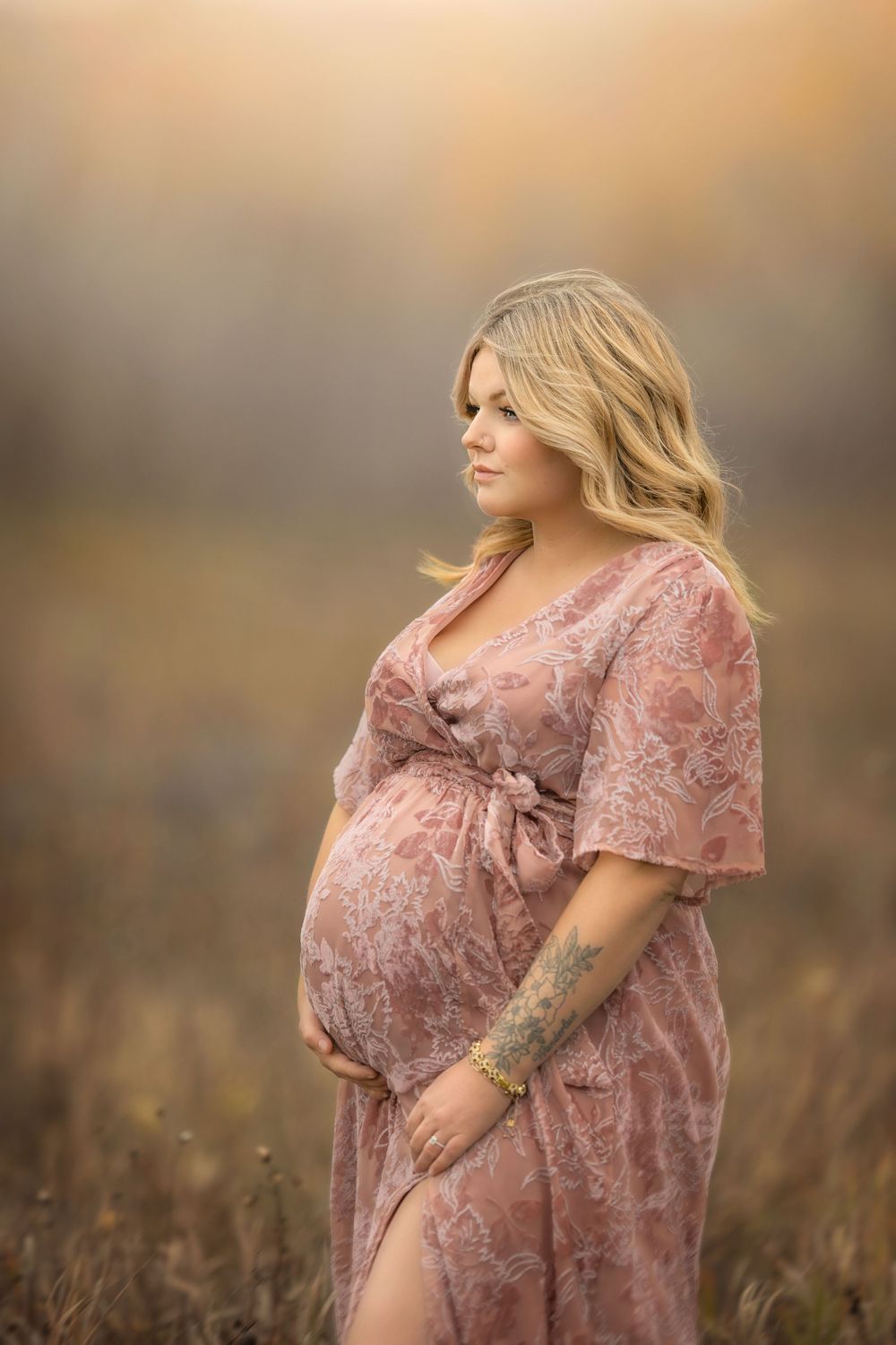 A glowing maternity portrait in a field at sunset featuring a pink velvet floral gown.