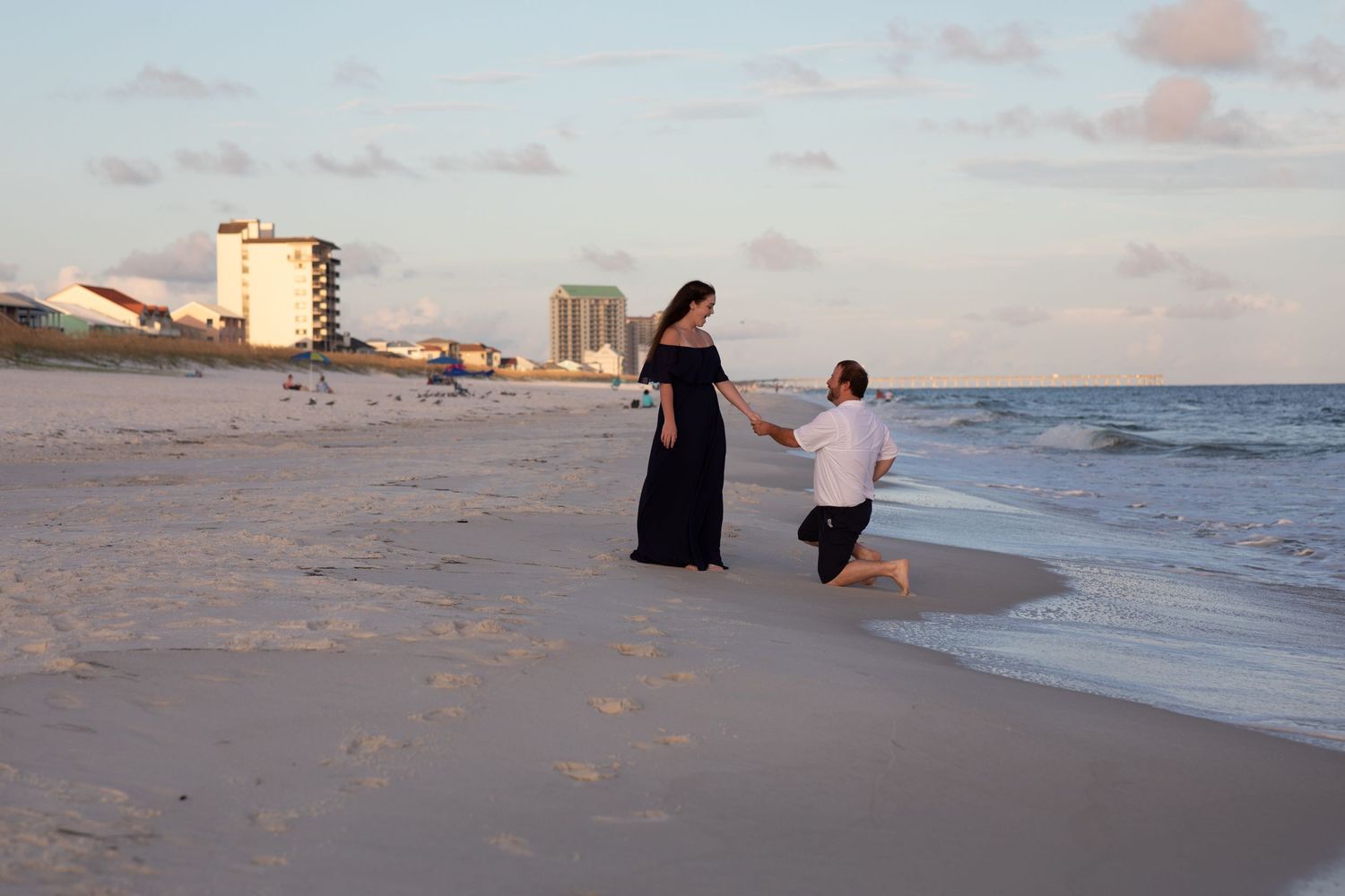 Sunset beach proposal moment captured with ocean waves and coastal architecture in background.