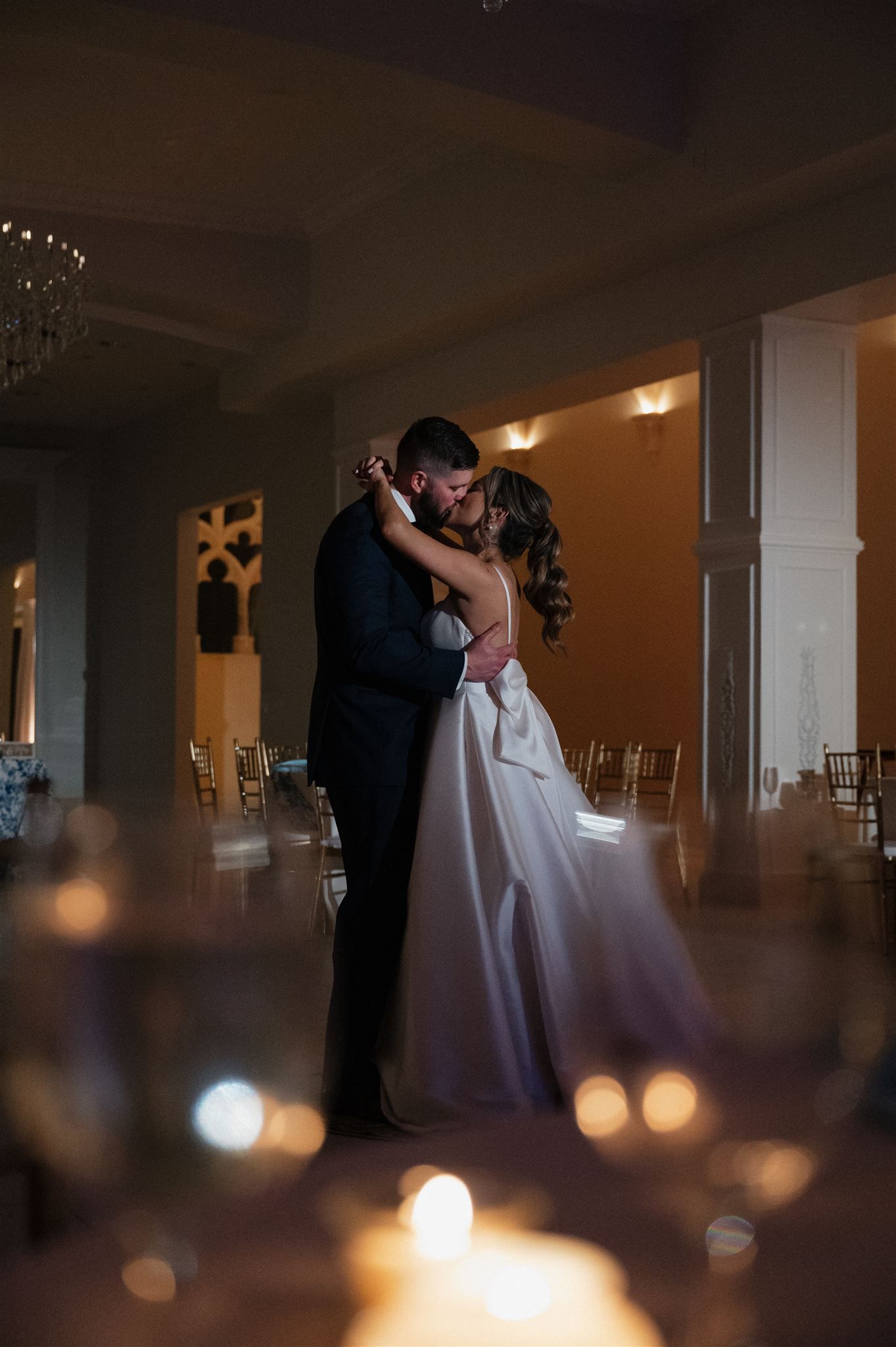 A romantic wedding couple dancing in a dimly lit ballroom with bokeh lights in the foreground.
