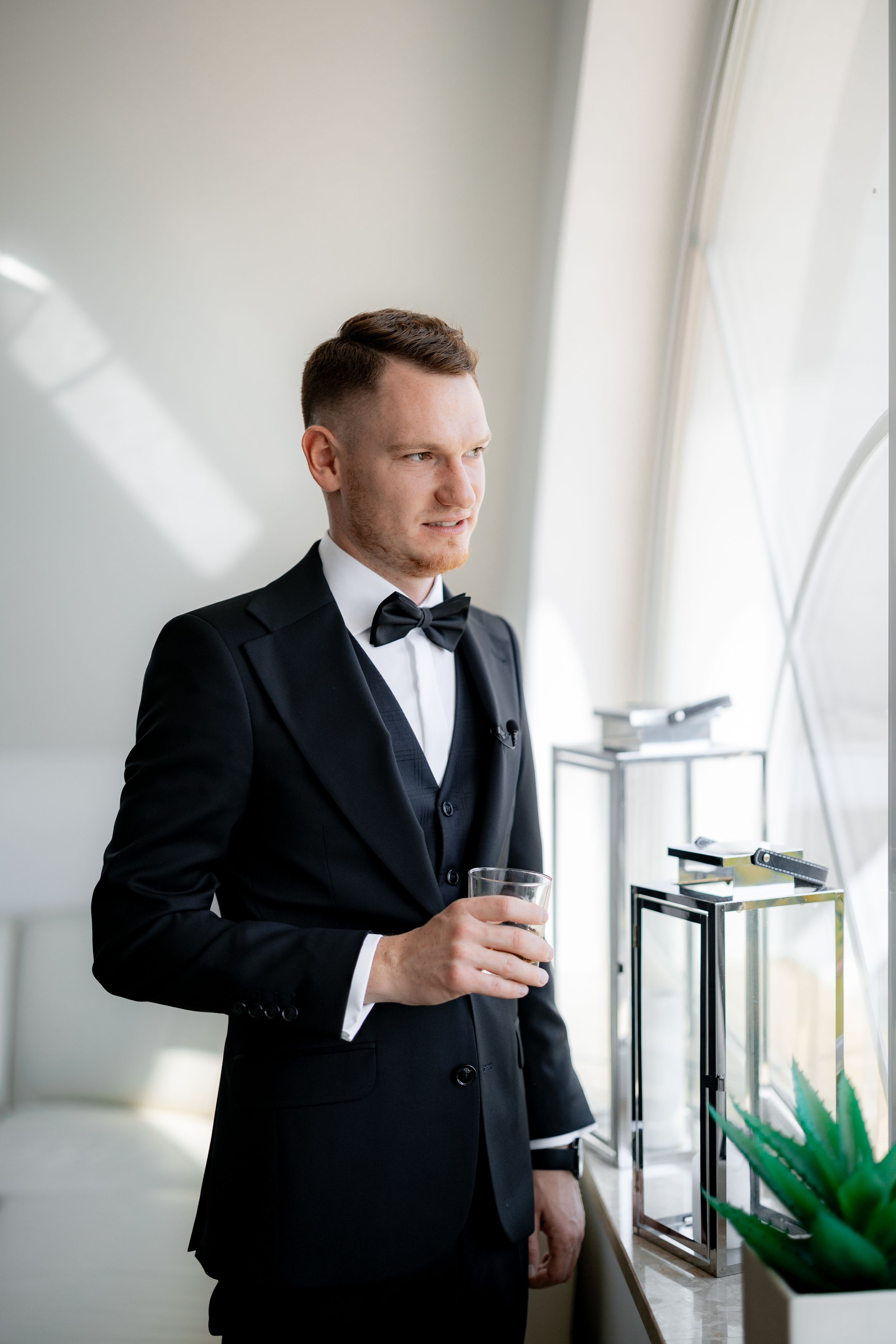 Groom in classic black tuxedo holding drink while standing by window in modern interior.