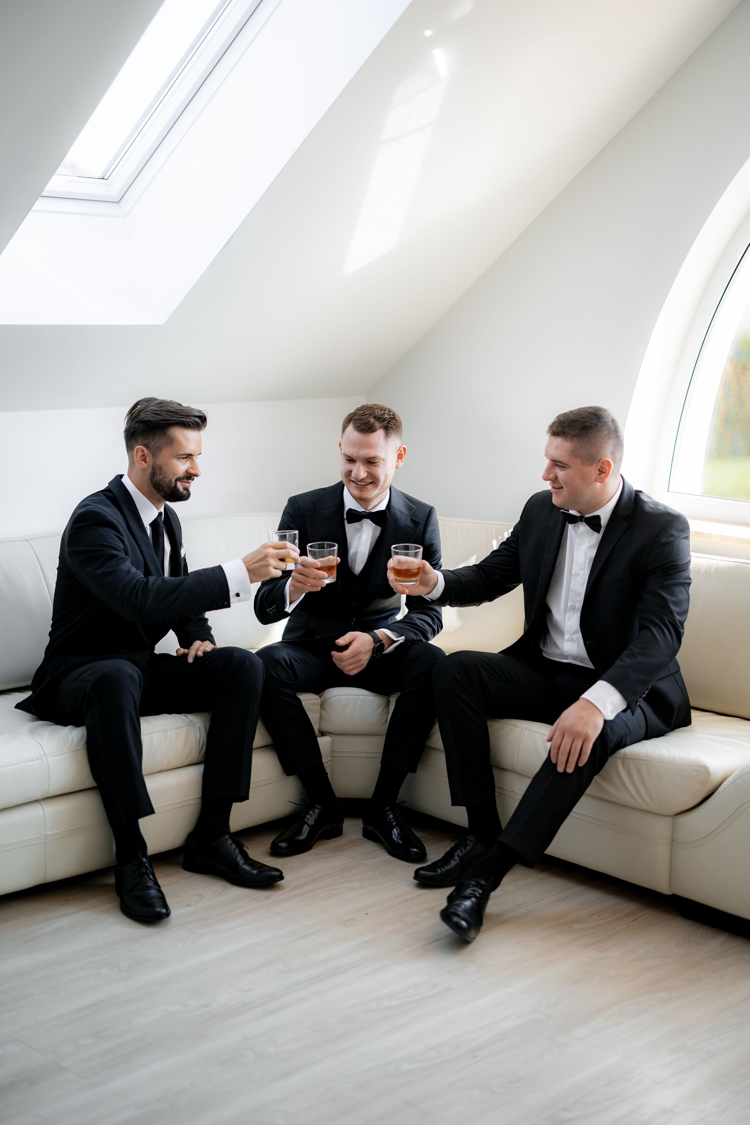 Three groomsmen in black tuxedos sharing drinks on a white couch under a skylight.