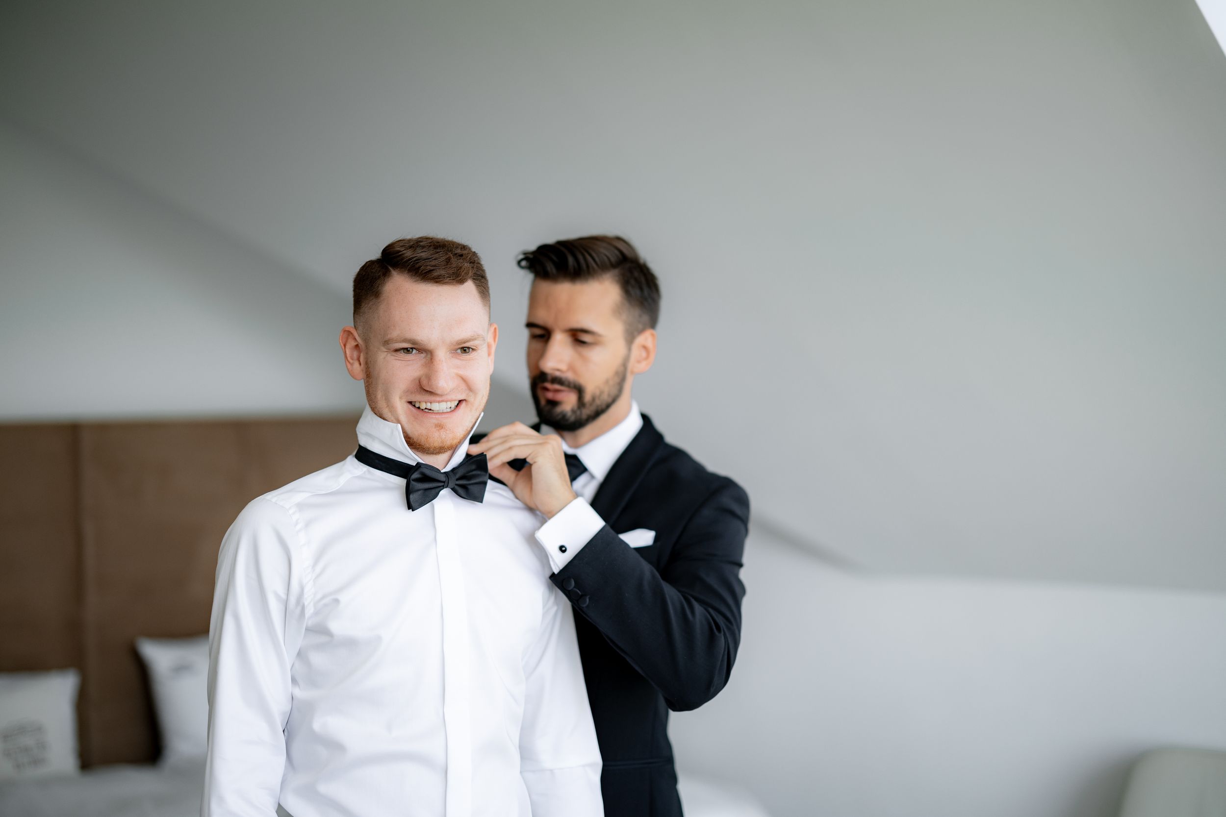 Two men in formal wear adjusting bow tie during wedding preparations.