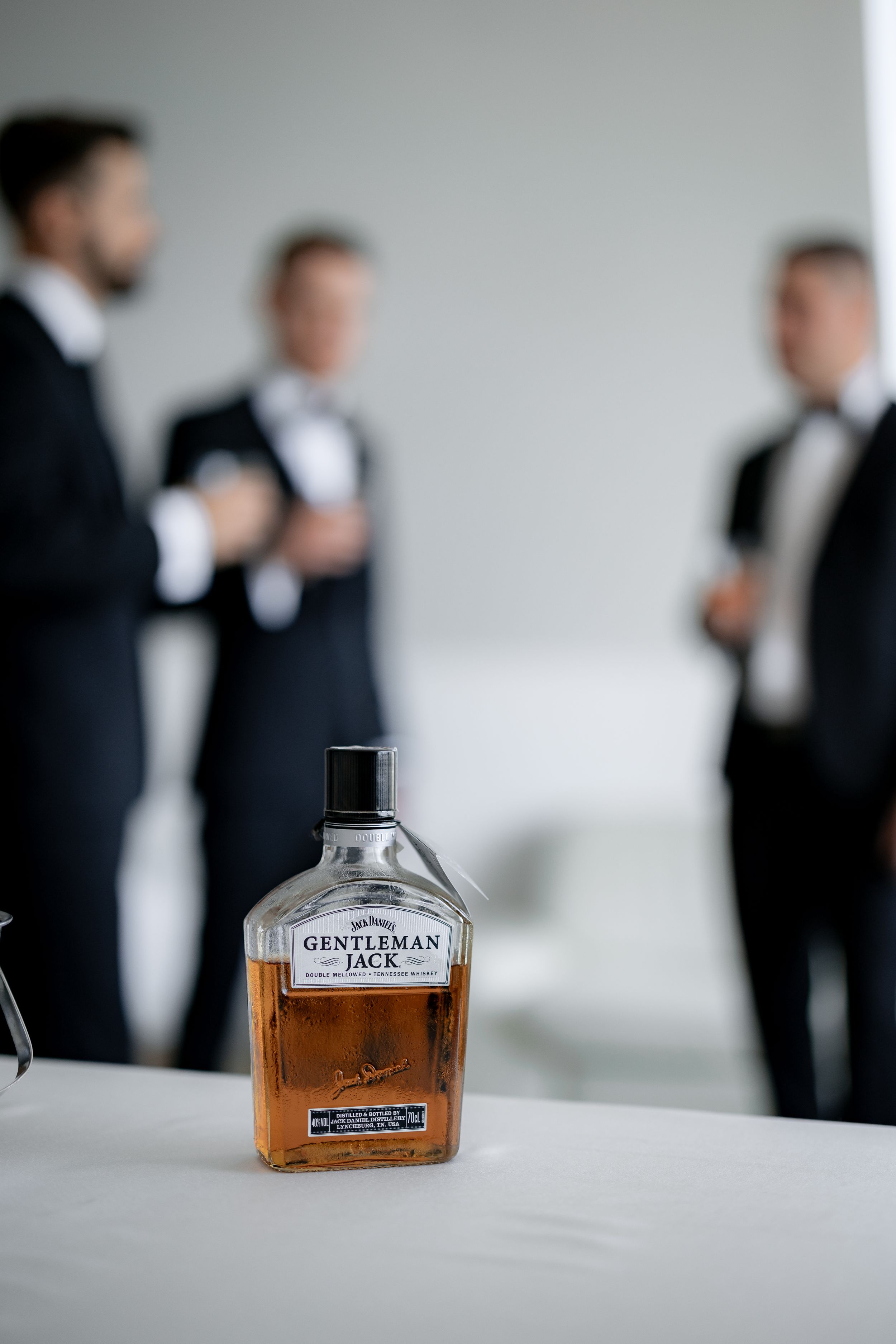 A bottle of Gentleman Jack whiskey sits on a table with figures in tuxedos blurred in background.