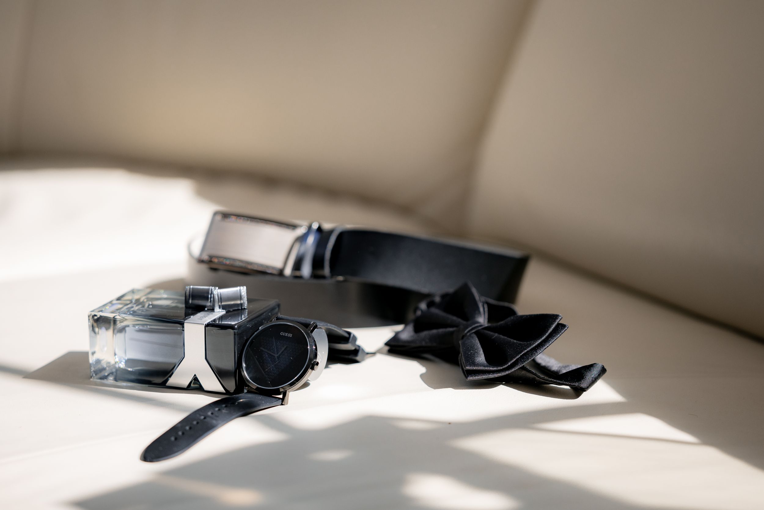 Groom's accessories including watch, bow tie and cufflinks arranged in sunlight.