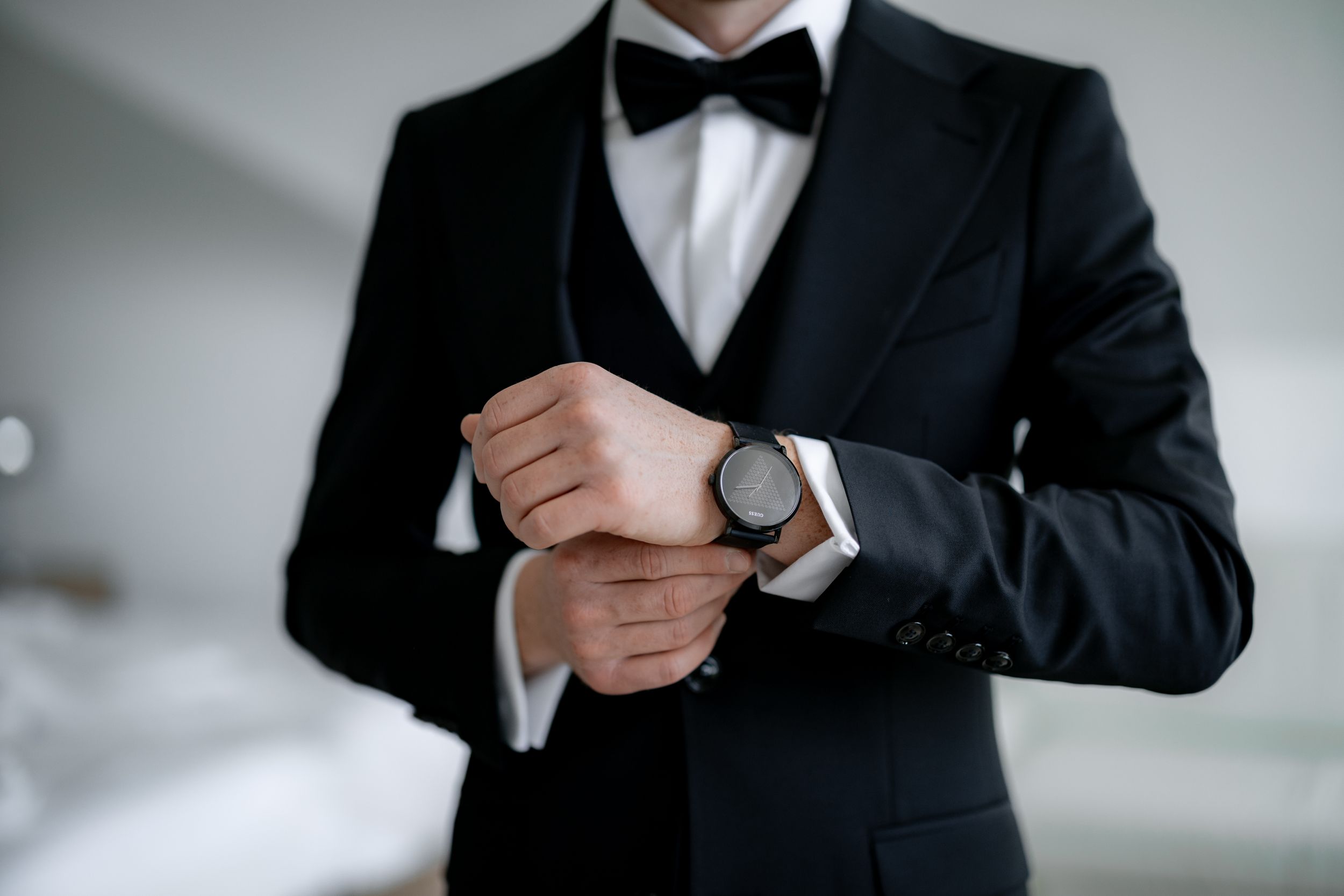 Close-up of a groom adjusting his silver watch while wearing a classic black tuxedo with bow tie.