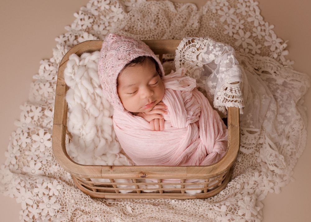 Sleeping newborn baby wrapped in pink swaddle lying in wooden basket surrounded by delicate lace.