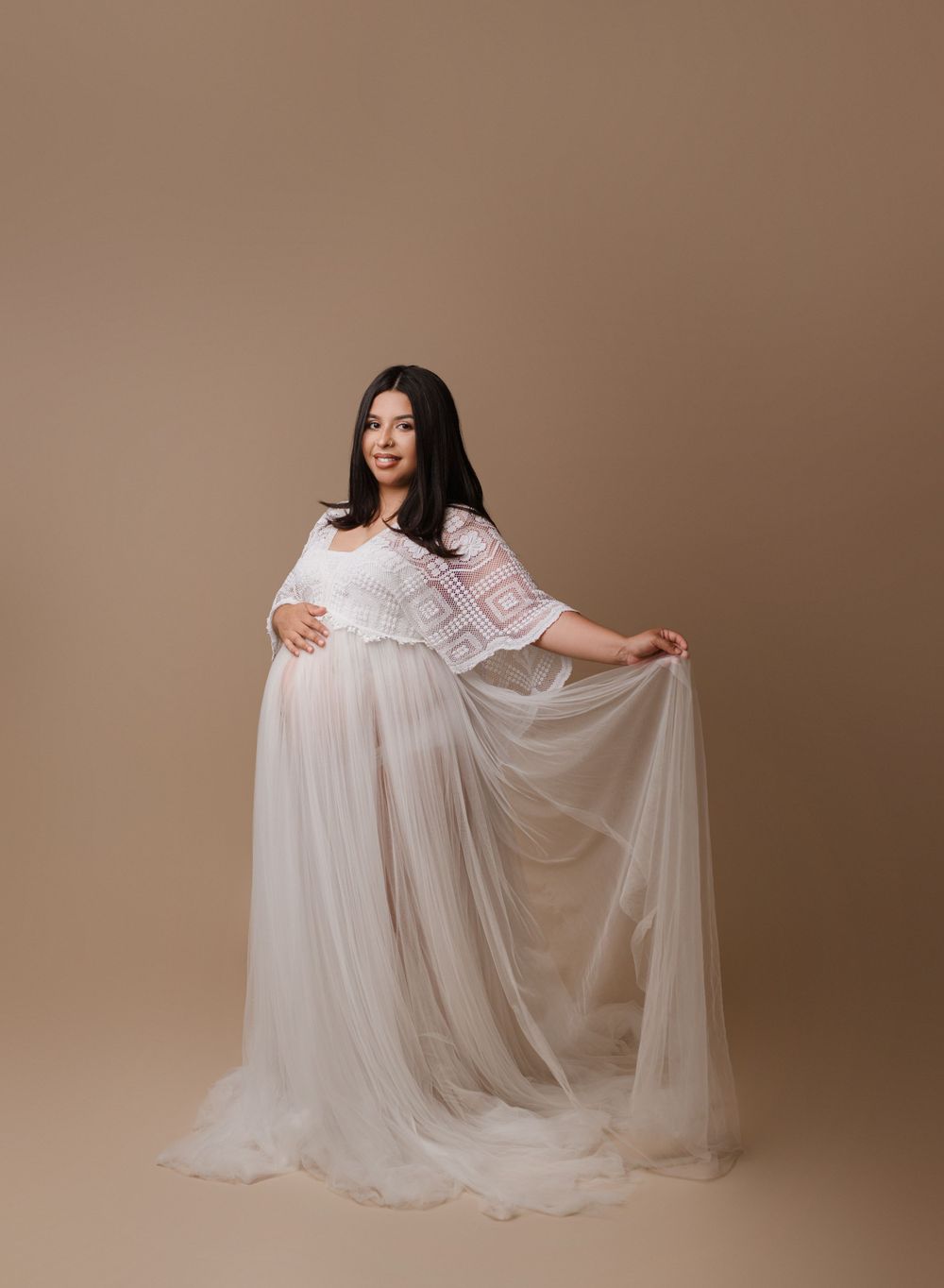 Expectant mother in flowing white maternity gown poses against beige backdrop.