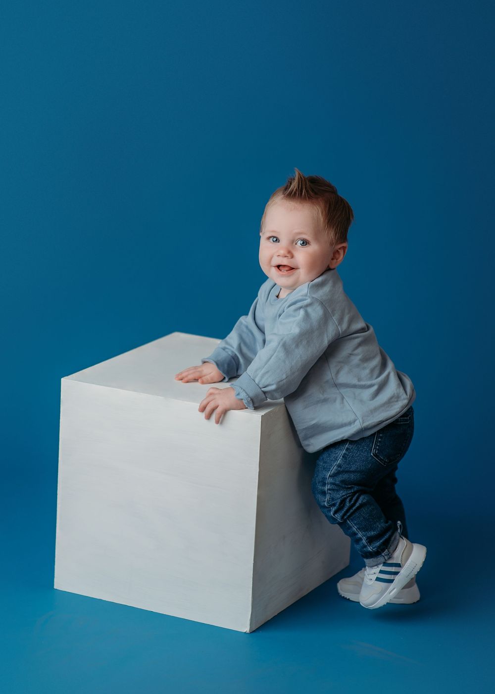 A joyful baby in blue clothing climbing on a white cube against a deep blue background.