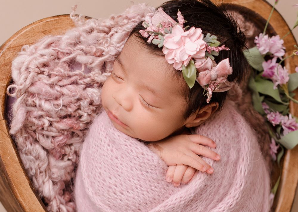 A sleeping newborn wrapped in pink with floral crown in wooden bowl surrounded by flowers.