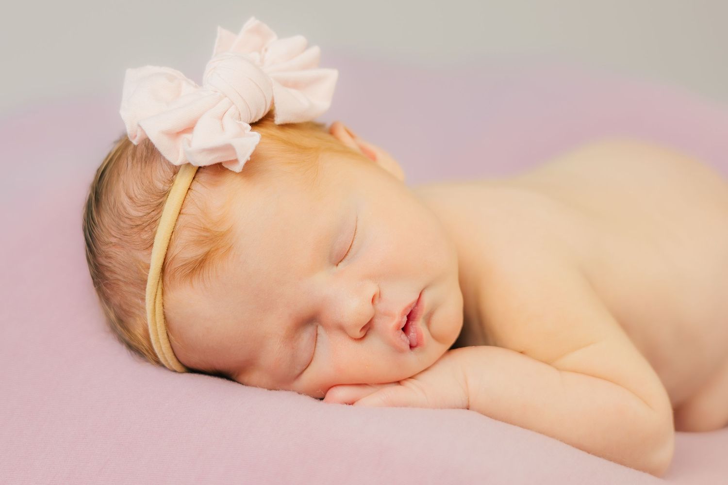 Sleeping newborn baby wearing a pink bow headband on pink background.