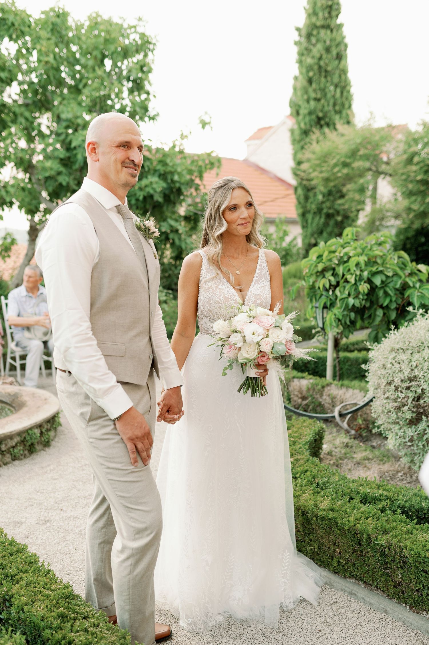 A couple posing in a Mediterranean garden setting wearing elegant wedding attire.