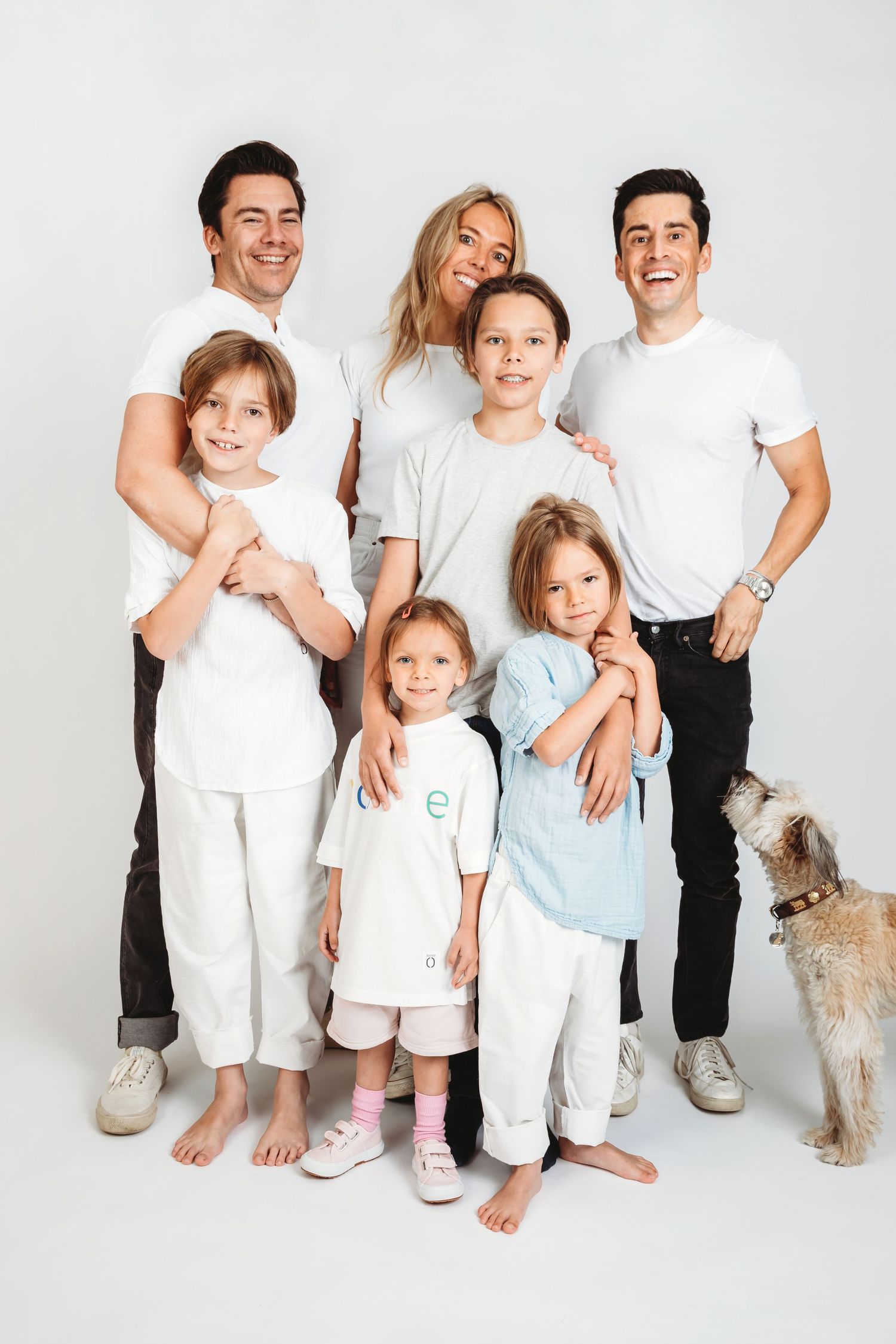 A casual family portrait in white outfits with four children and a small dog against a white background.