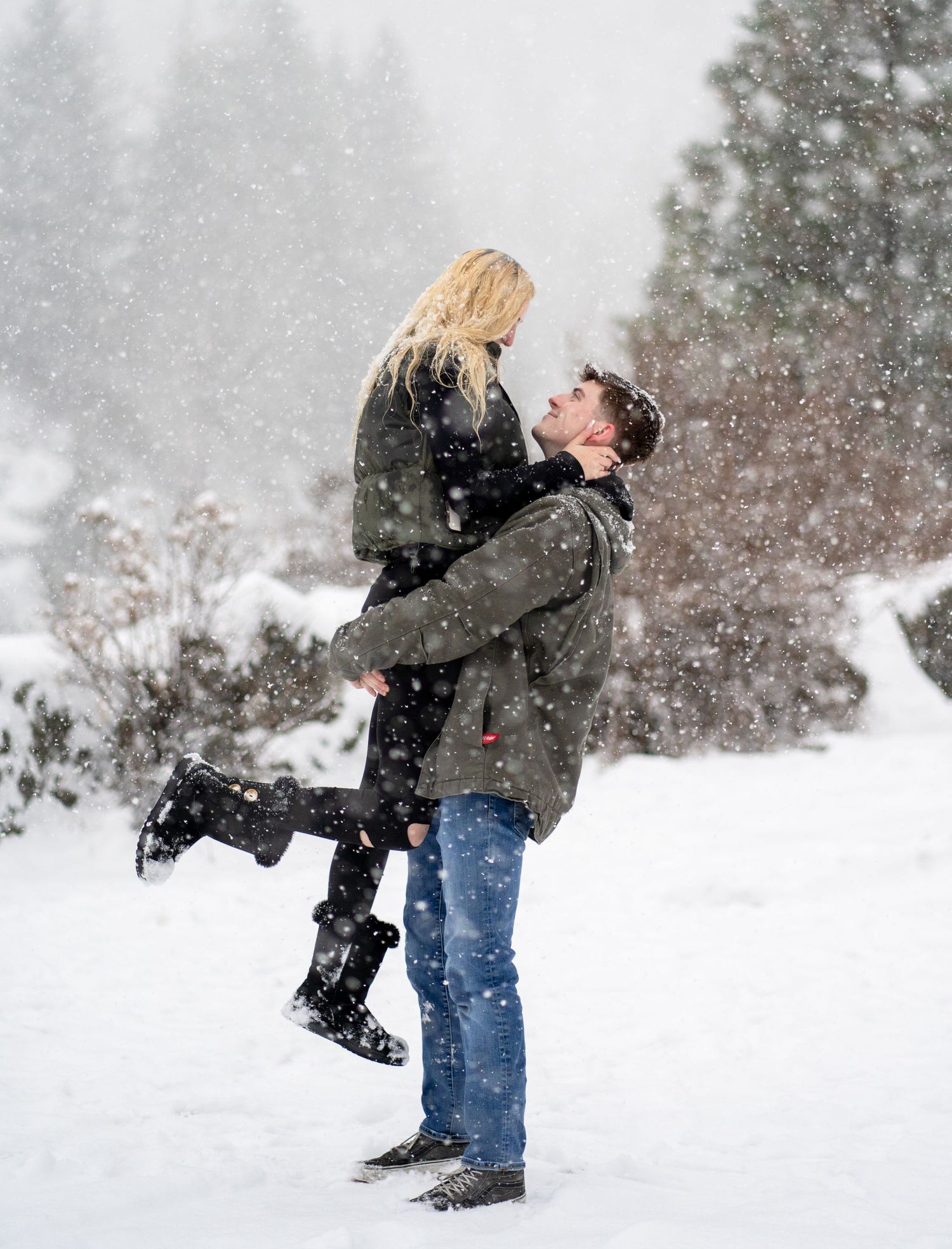 A joyful lift pose during a snowy winter photoshoot with pine trees in the background.