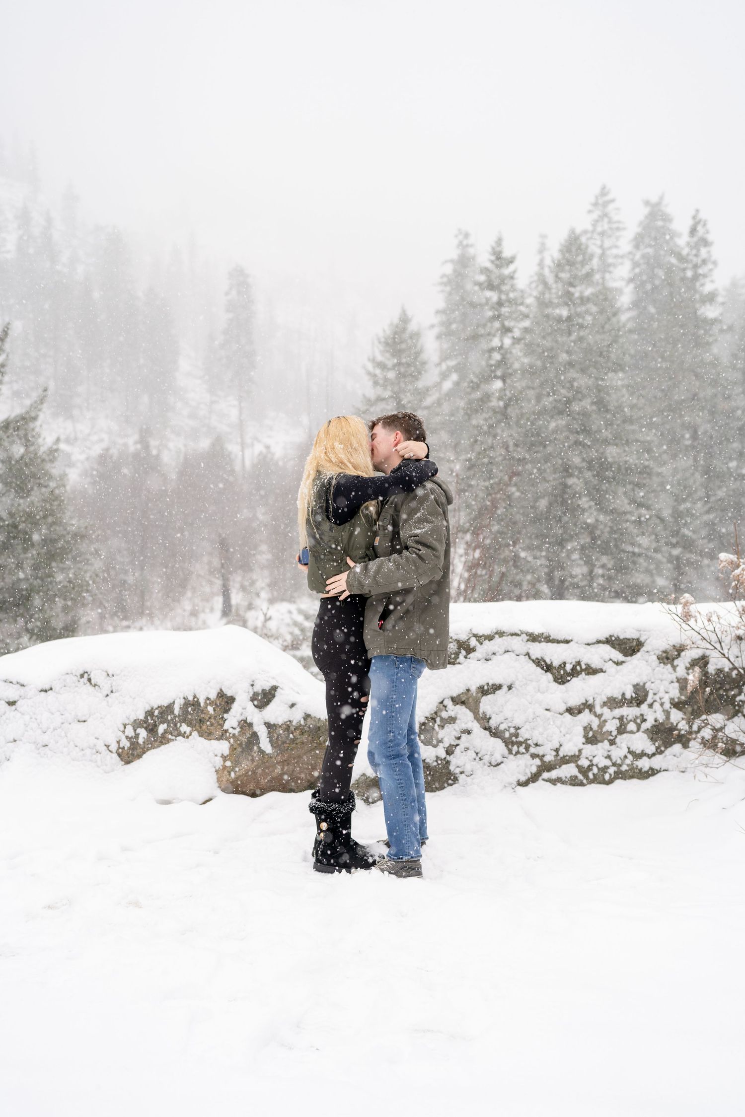 A couple shares an intimate moment in the falling snow with evergreen trees behind them.