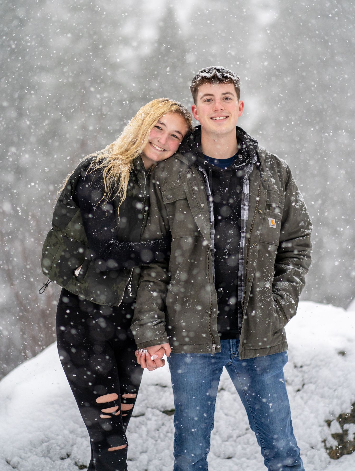 A happy couple poses together in winter coats while snow falls in a forest setting.