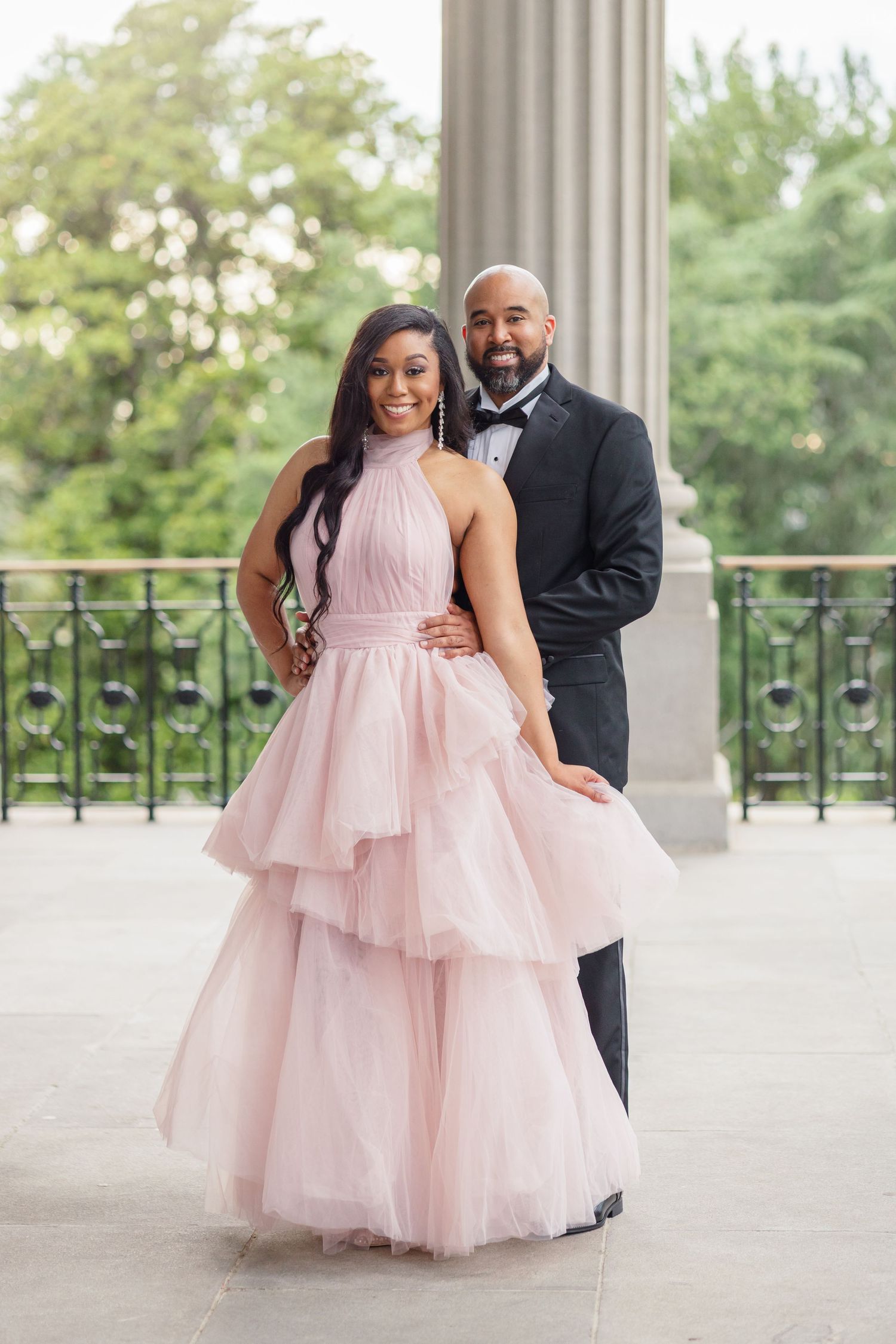 A couple poses elegantly on a balcony, with the woman wearing a pink tulle gown and the man in a black tuxedo.