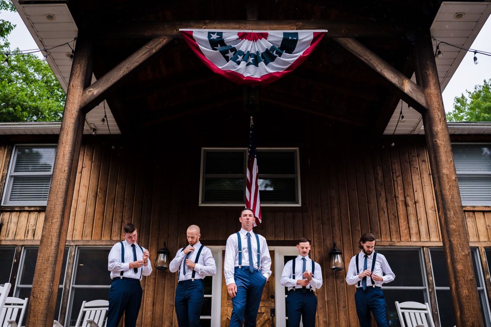 Wedding party members in suspenders and formal attire lined up under a rustic wooden pavilion.