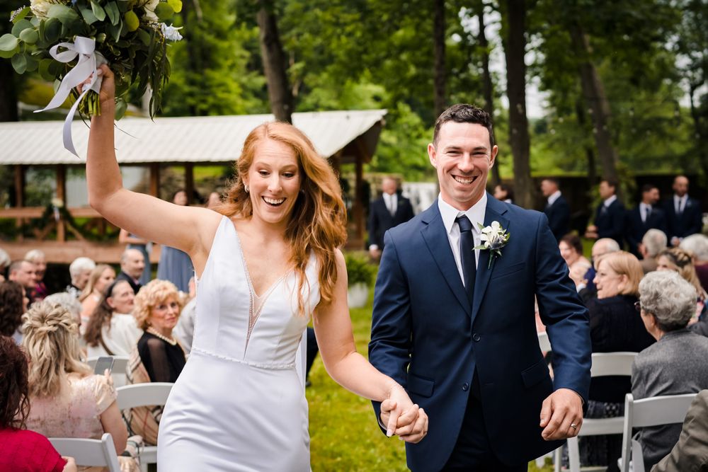 Outdoor wedding ceremony setup with white chairs and floral arrangements in a wooded setting.