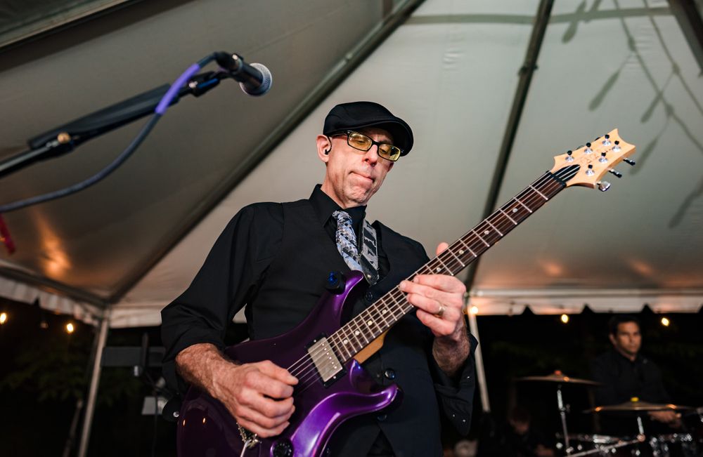 Guitarist in formal attire plays purple electric guitar during evening performance.