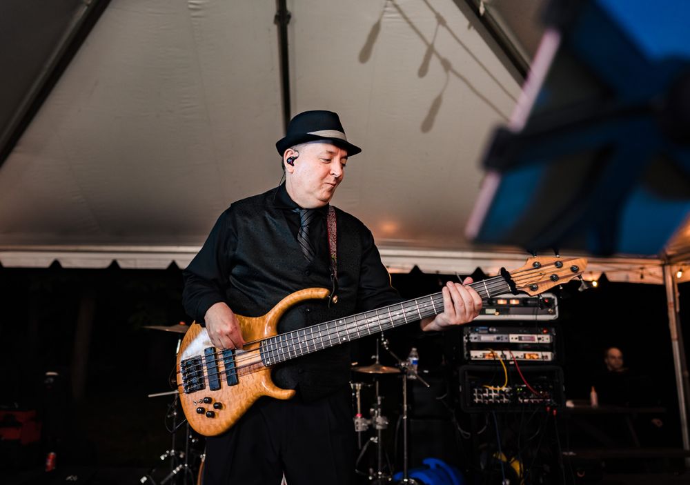 Bass guitarist in black attire and hat performs on stage under tent lighting.