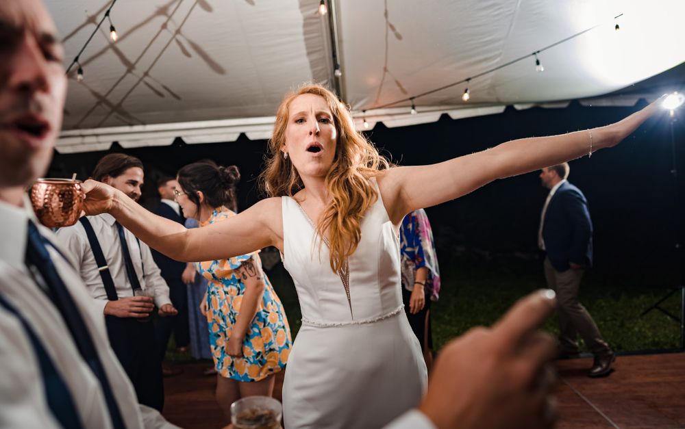 Dancing guest in white dress spreads arms wide during reception celebration.