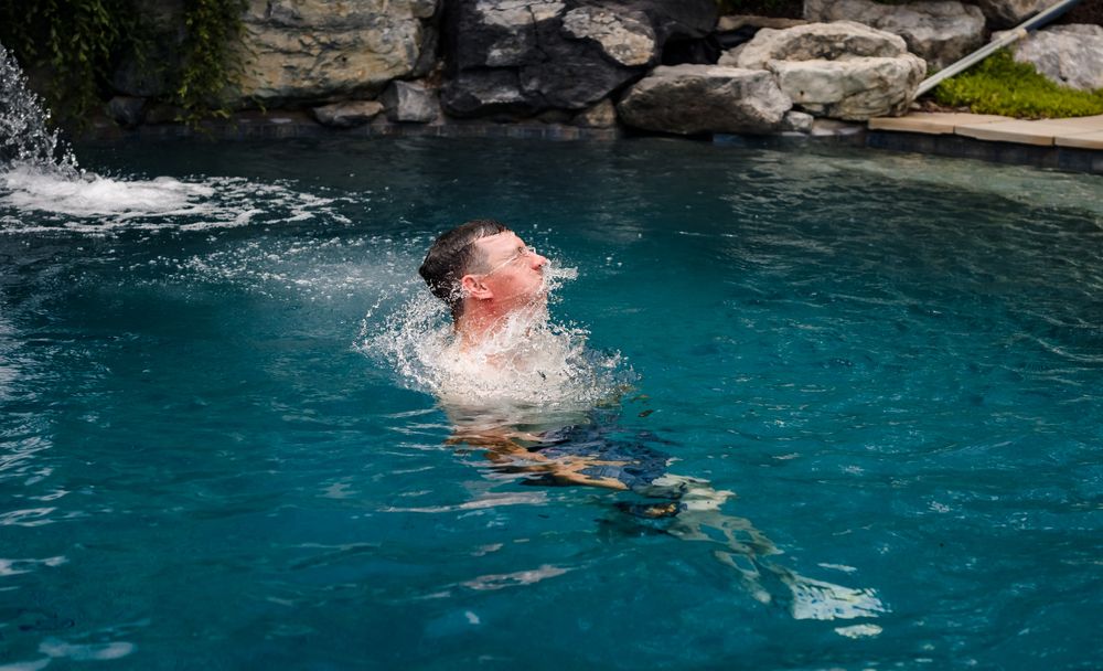 Person swimming and splashing in a blue pool surrounded by rocky landscaping.