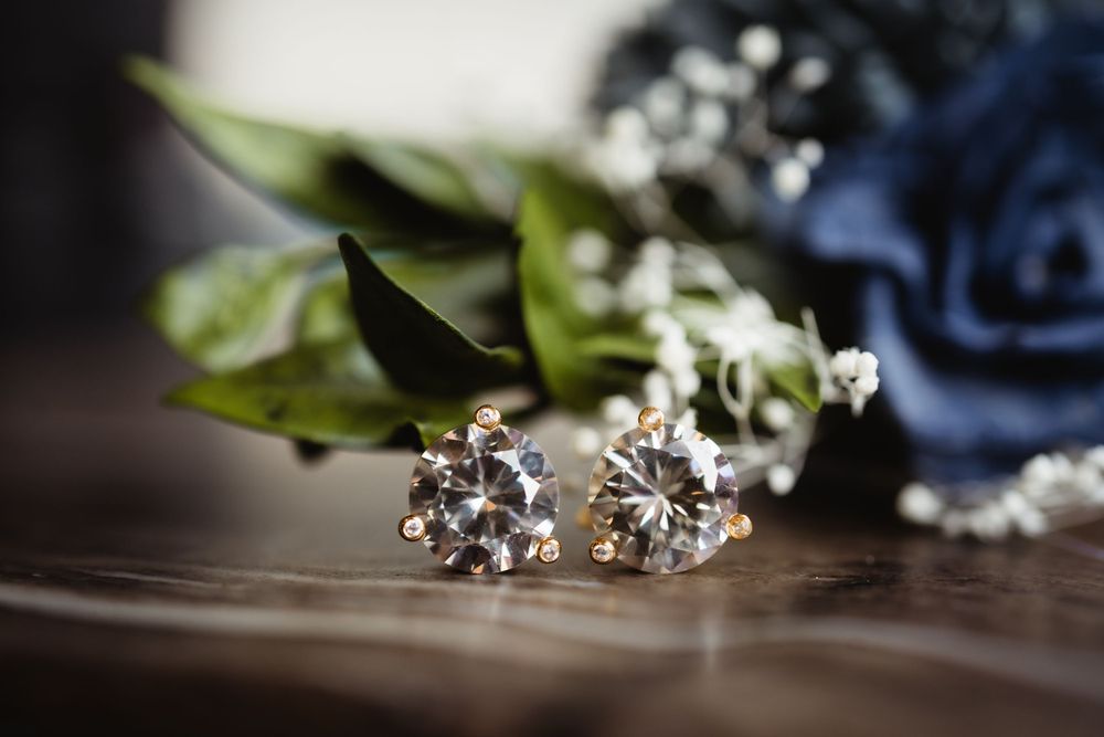 Diamond stud earrings displayed with blue flowers and greenery on wooden surface.