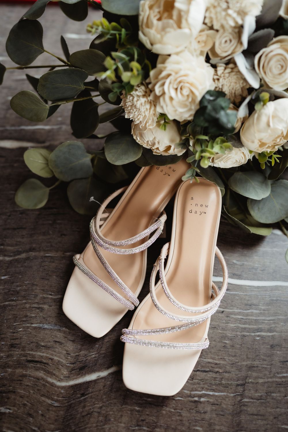 Crystal-embellished bridal sandals displayed with white roses and eucalyptus leaves.