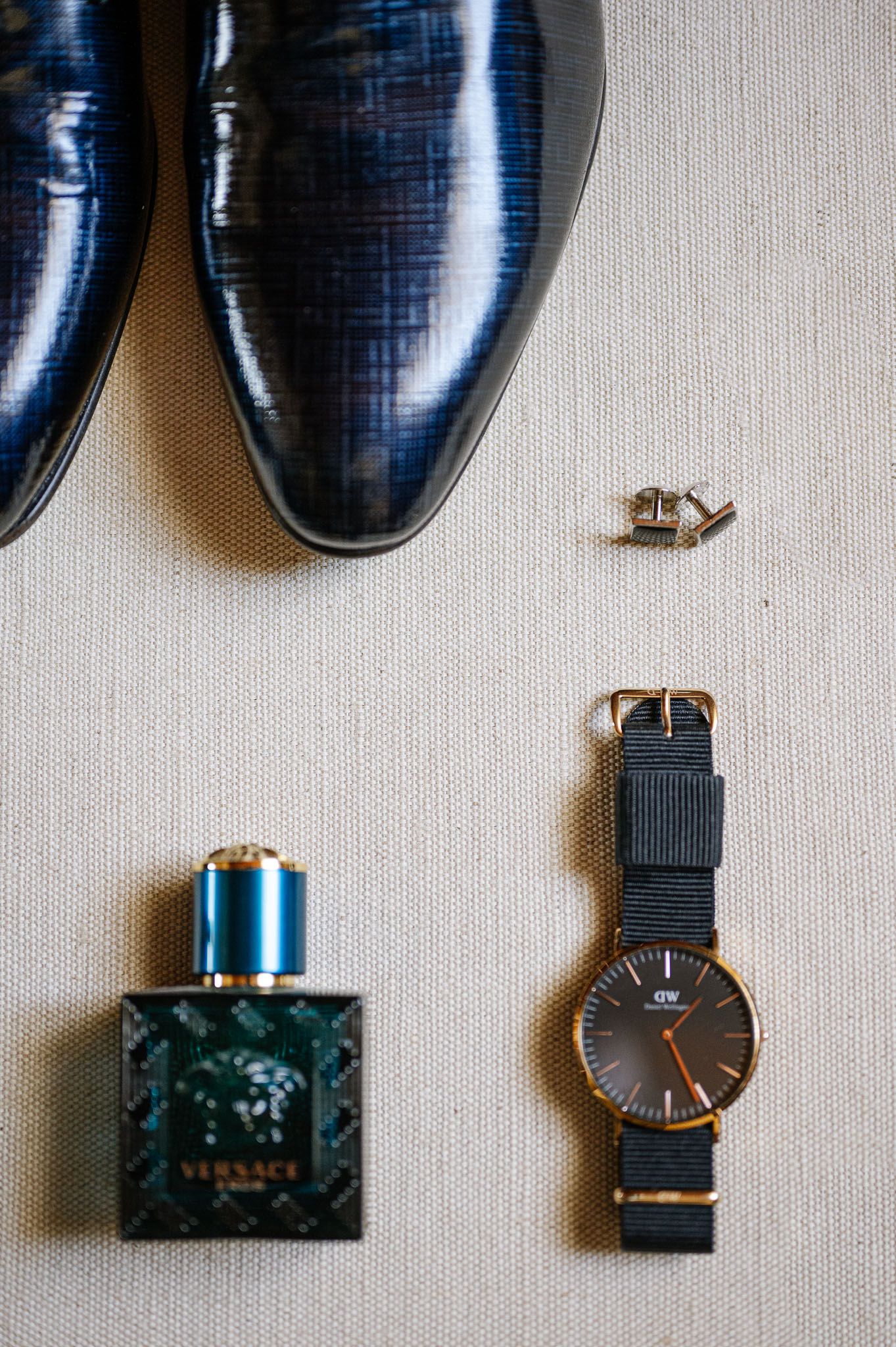 Wedding accessories including navy dress shoes, a wristwatch, and cologne bottle arranged on light fabric.