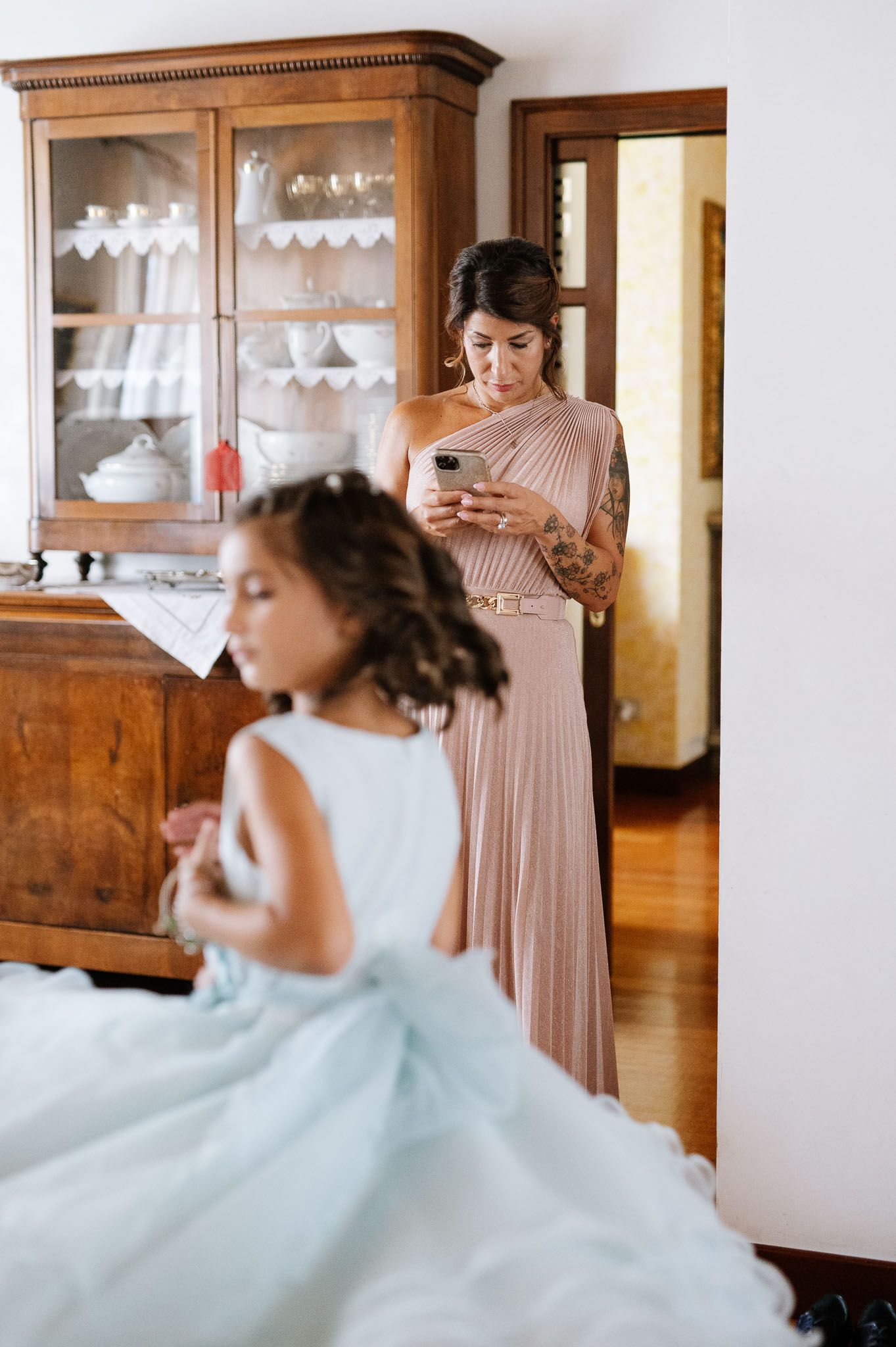 Wedding preparations showing bridesmaid in pink dress and flower girl in blue tulle dress near antique cabinet.