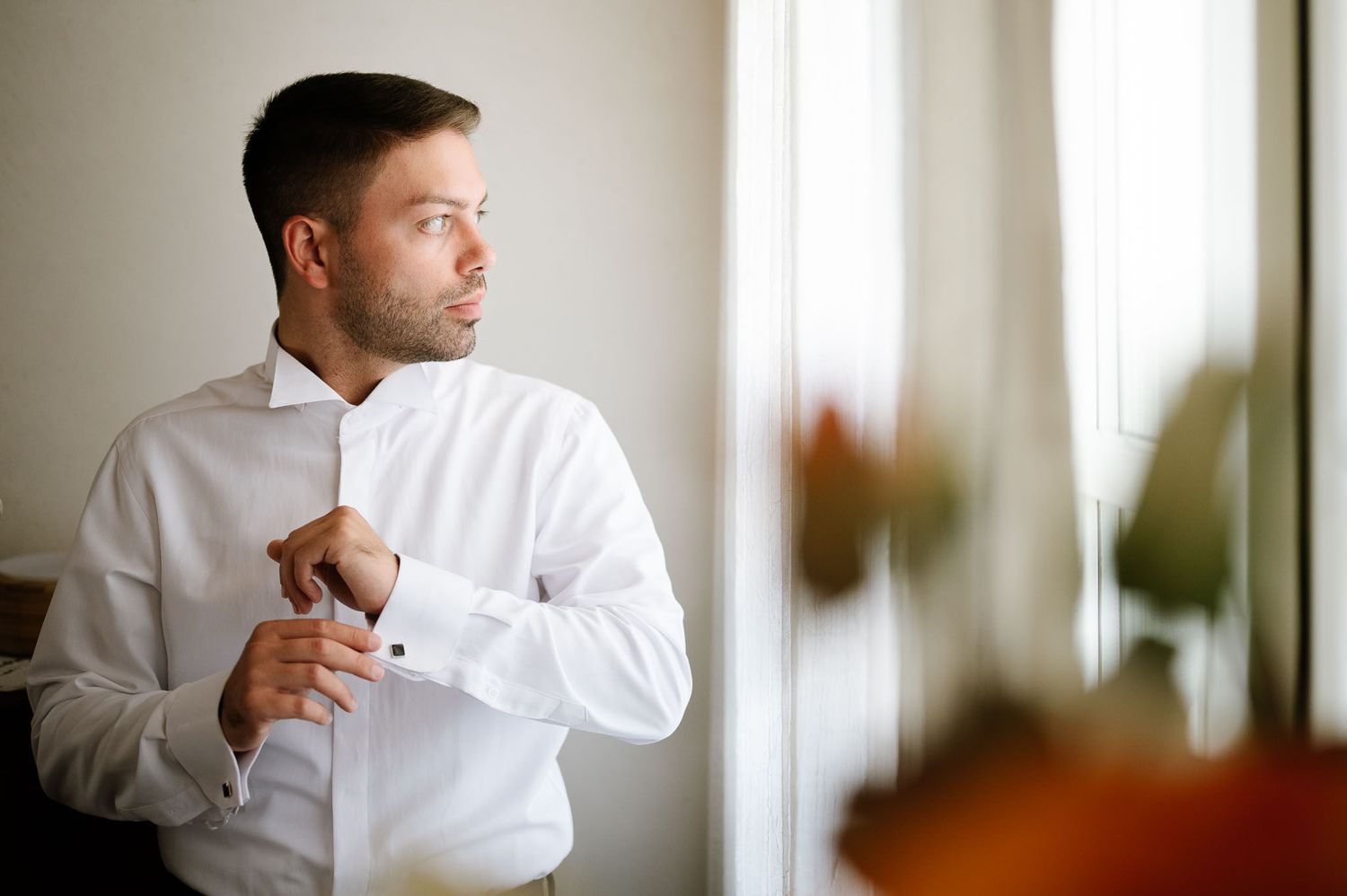 Person in white dress shirt adjusts cuffs while looking thoughtfully out window.