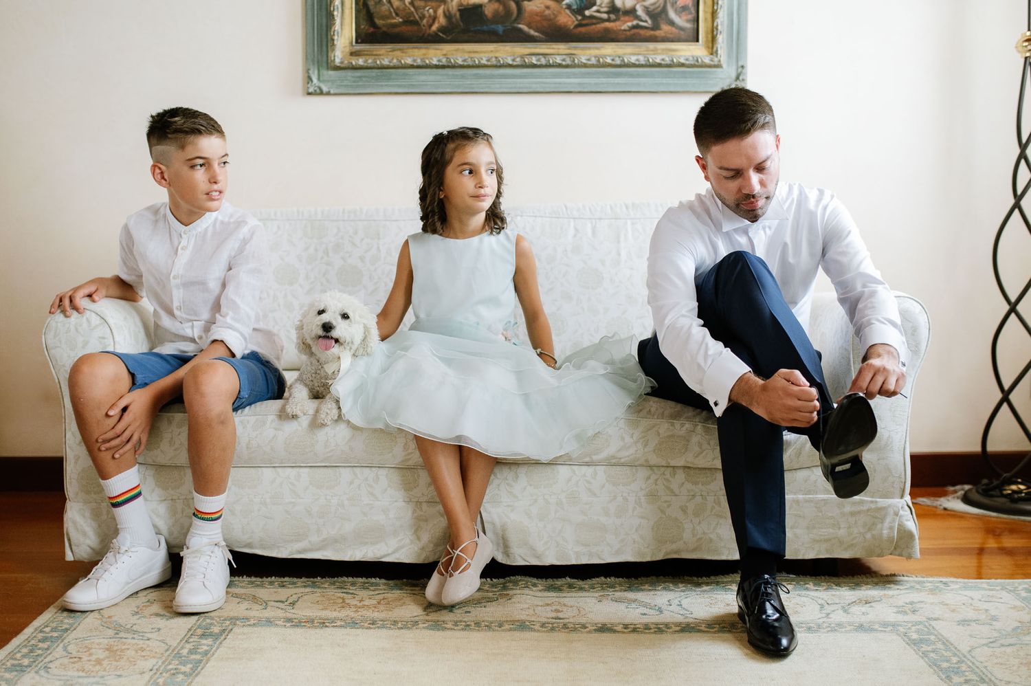Young boy, girl in white dress, and adult sit on white couch with small white dog.