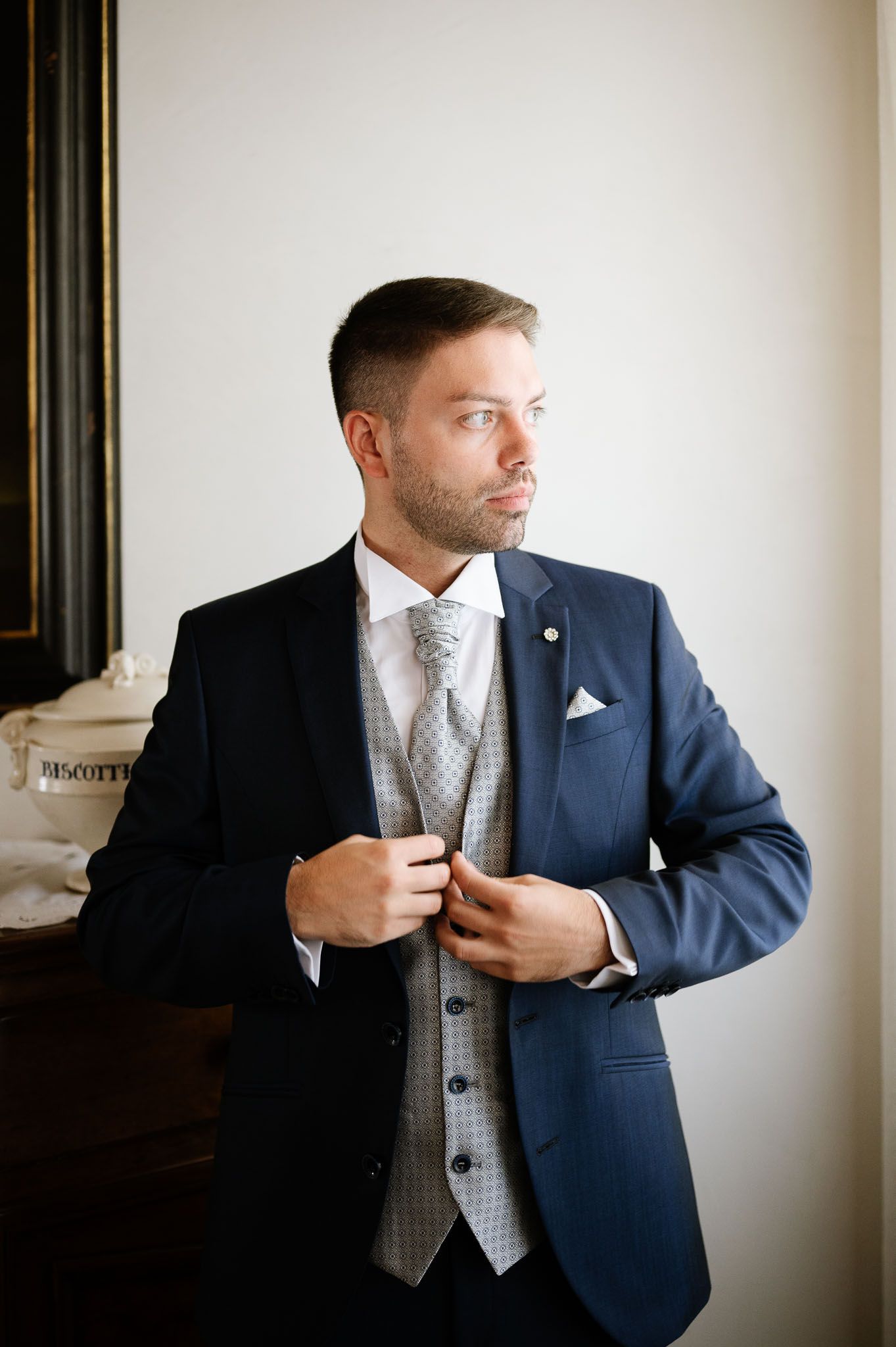 Groom in navy suit with grey waistcoat adjusts jacket in formal portrait.