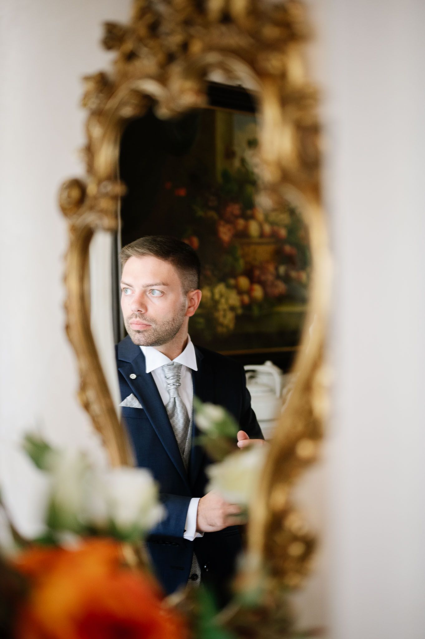 Reflection of groom in ornate gold mirror wearing navy suit and grey tie.
