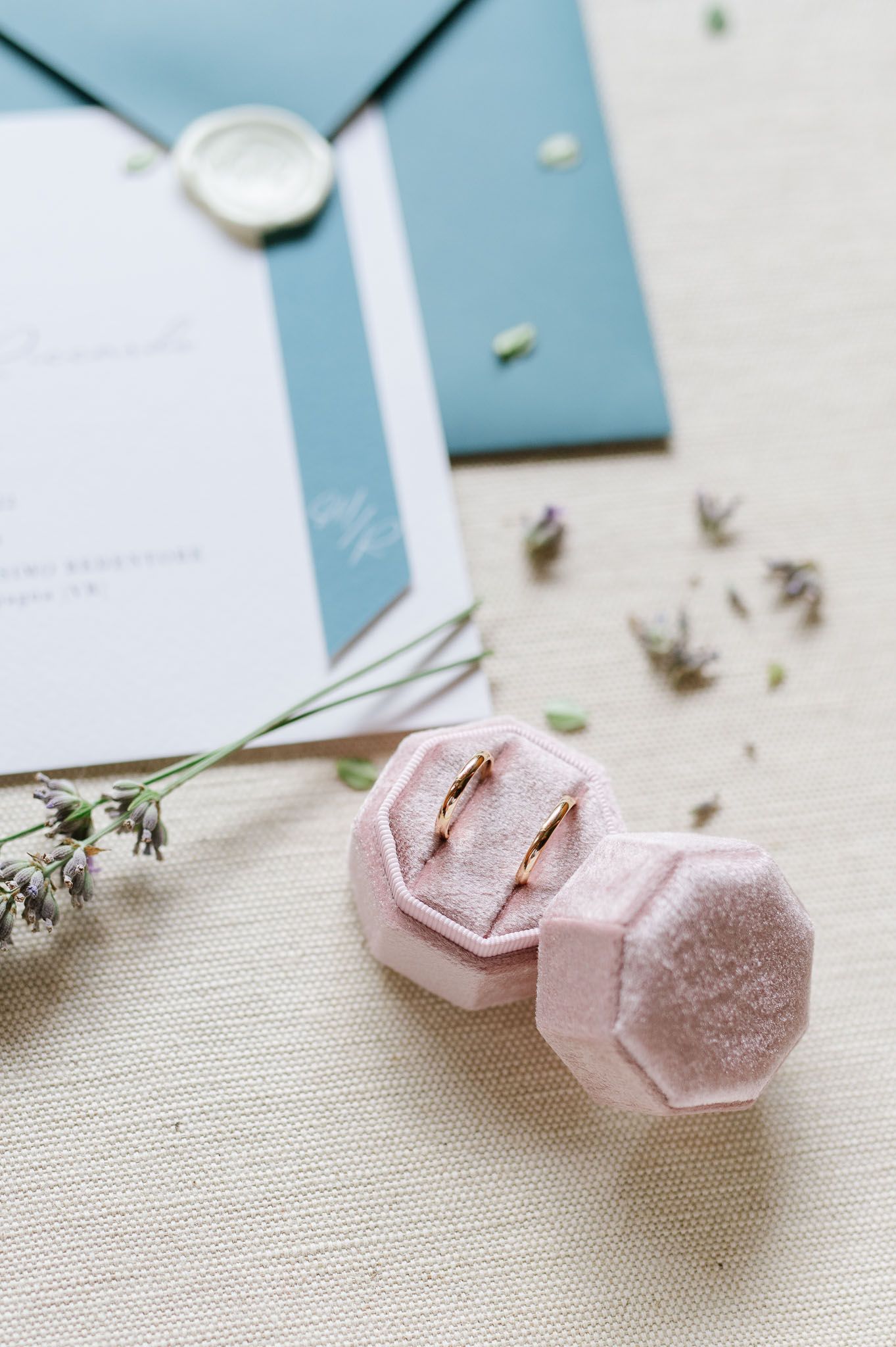 Wedding rings in pink velvet box with blue envelope and dried flowers on linen background.