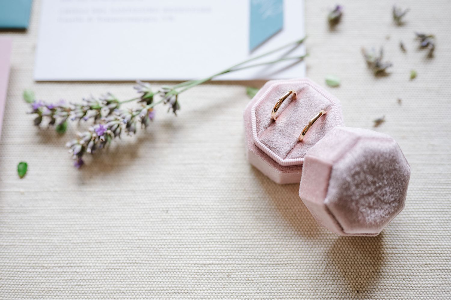 Pink velvet ring box with wedding rings and dried lavender on white linen surface.