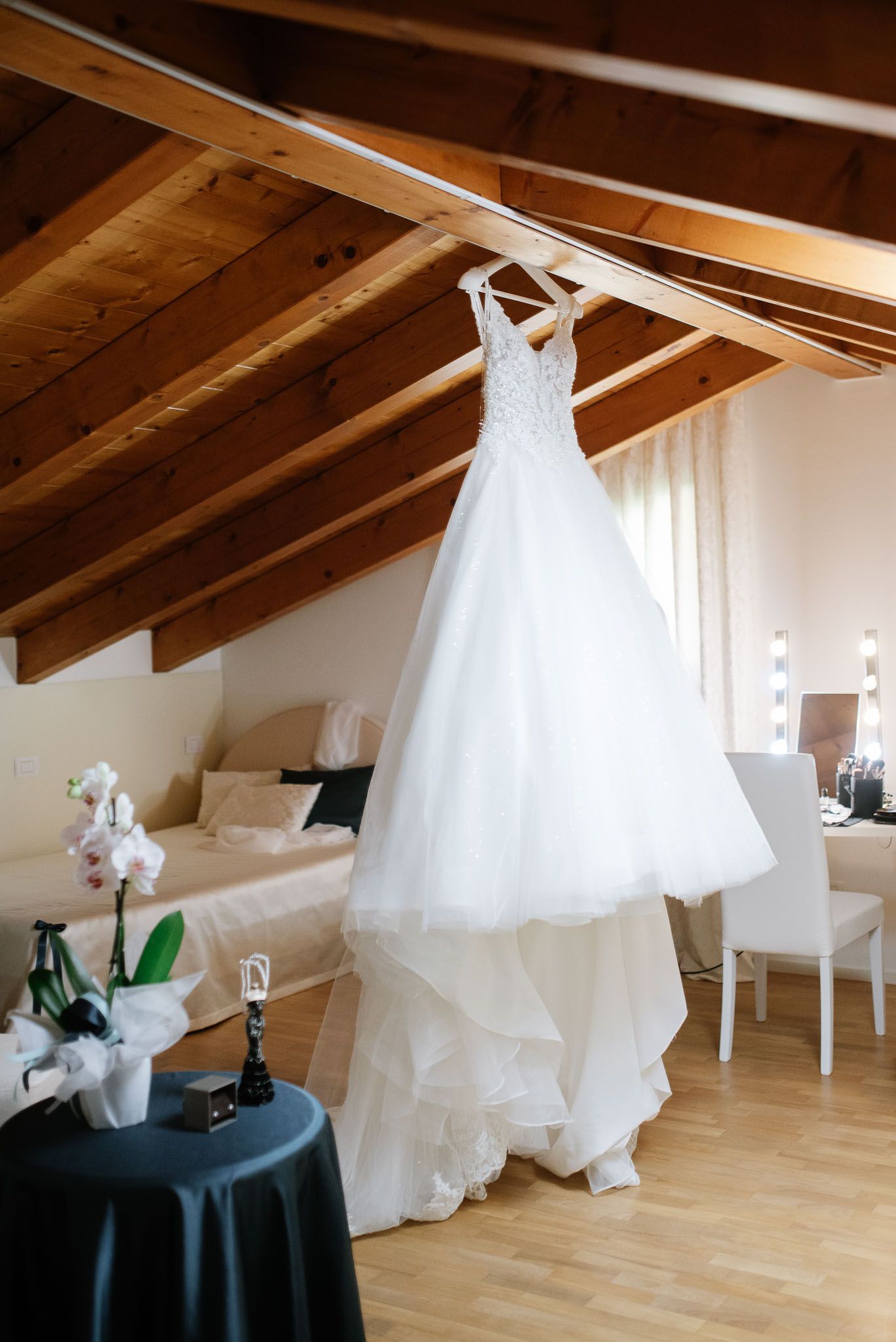 A white tulle wedding dress hangs from wooden beams in a rustic room with natural lighting.