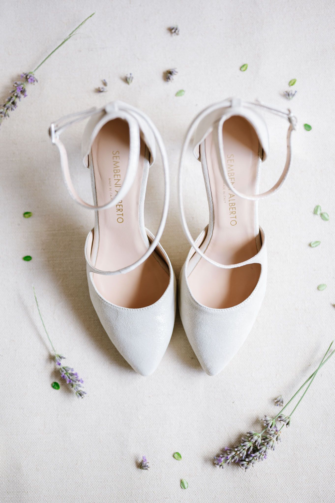 Elegant white bridal flats with ankle straps displayed with sprigs of lavender.