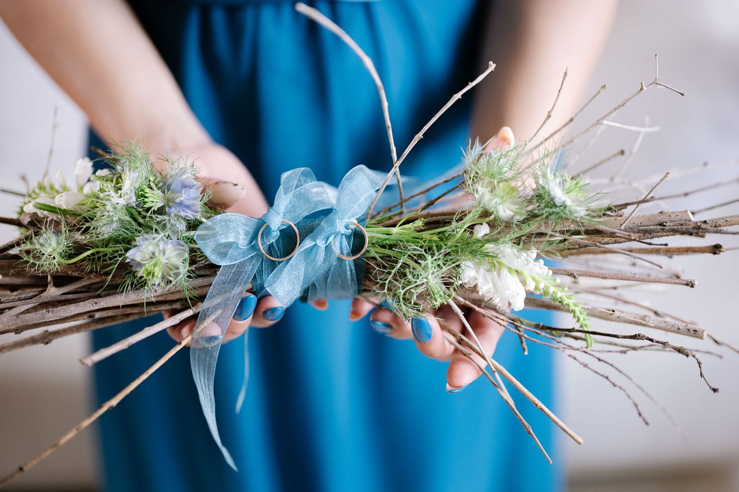 Hands holding a blue floral arrangement with twigs and small flowers against a blue background.